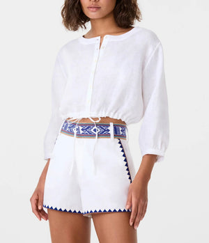 Woman wearing an Emporio Sirenuse New Jinny Blue Horizon Top, a white linen cropped style with blue embroidered patterns, paired with high-waisted white shorts featuring matching blue embroidery.
