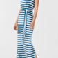 A woman wears the Emporio Sirenuse Kimi Ikat Stripes Dress, a sleeveless blue-and-white handwoven maxi with a matching waist tie, paired with brown flat sandals.