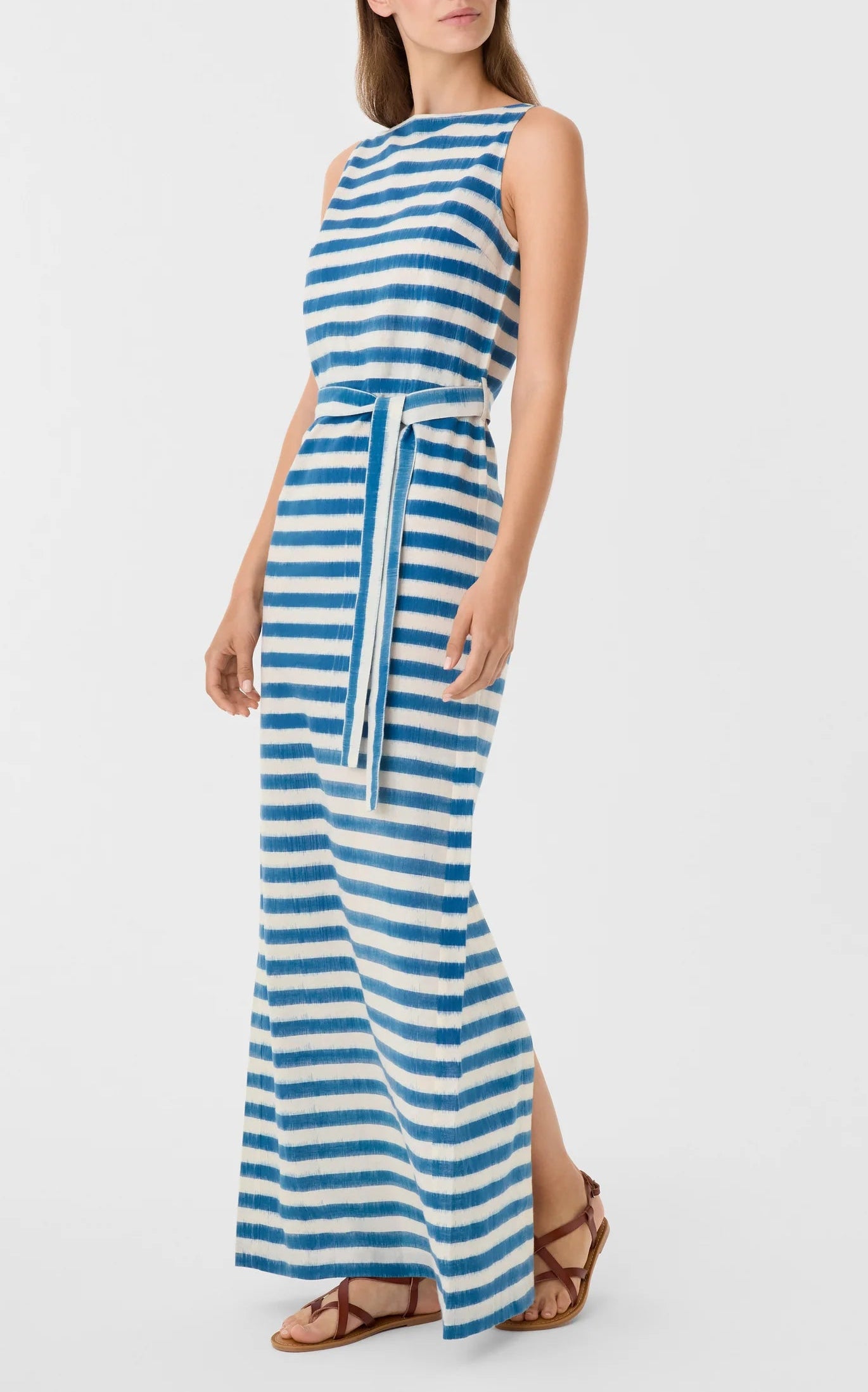 A woman wears the Emporio Sirenuse Kimi Ikat Stripes Dress, a sleeveless blue-and-white handwoven maxi with a matching waist tie, paired with brown flat sandals.