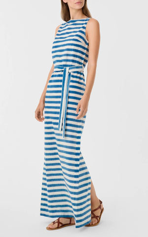 A woman wears the Emporio Sirenuse Kimi Ikat Stripes Dress, a sleeveless blue-and-white handwoven maxi with a matching waist tie, paired with brown flat sandals.
