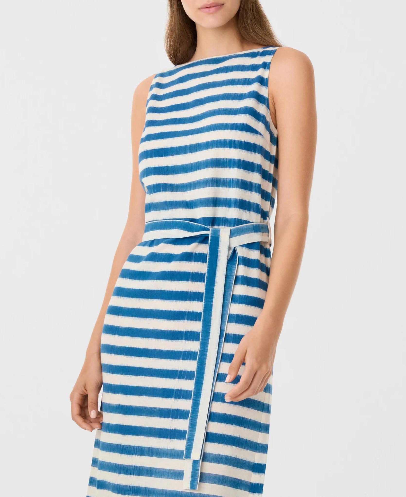 A woman wears the Emporio Sirenuse Kimi Ikat Stripes Dress, a sleeveless handwoven piece with horizontal blue and white stripes and a matching waist belt, photographed from the knees up against a plain background.