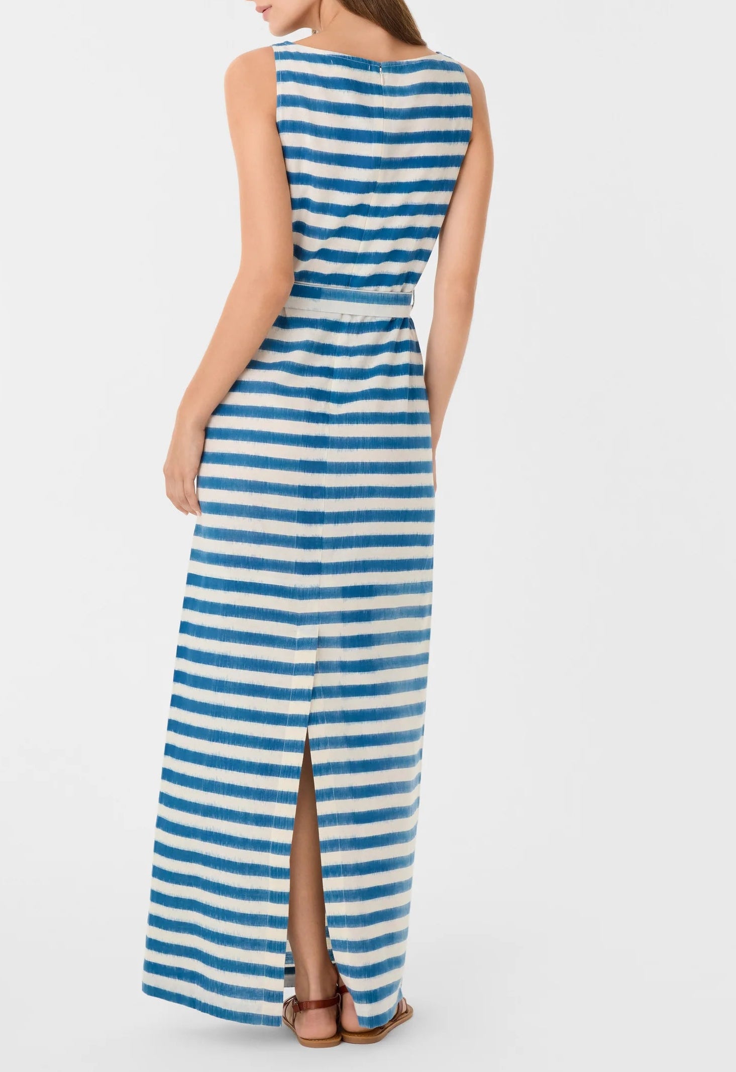 A woman wearing the Emporio Sirenuse Kimi Ikat Stripes Dress, a sleeveless blue and white handwoven maxi with a back slit, stands facing away from the camera.