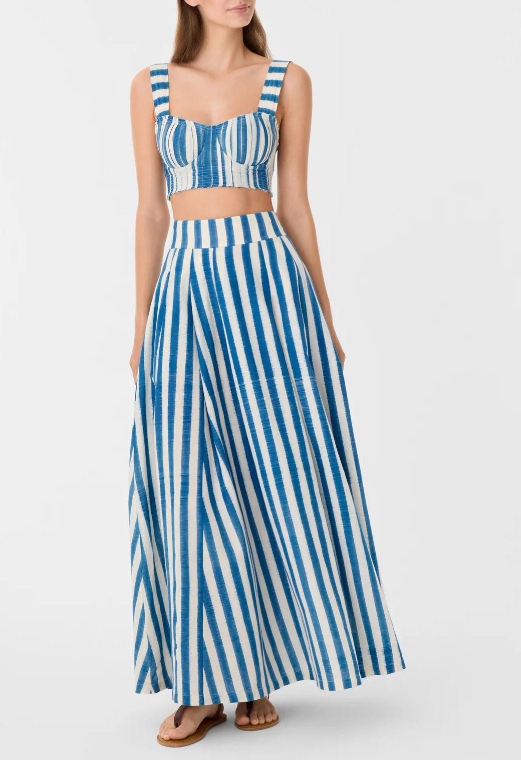 A woman wears the Emporio Sirenuse Flaminia Ikat Stripes Skirt with a matching blue and white crop top and sandals, her hands in her pockets.