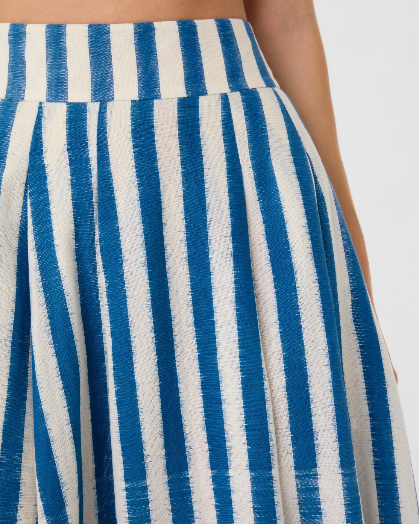 Close-up of a person wearing the Emporio Sirenuse Flaminia Ikat Stripes Skirt, crafted from handwoven pleated fabric with vertical blue and white Ikat stripes.