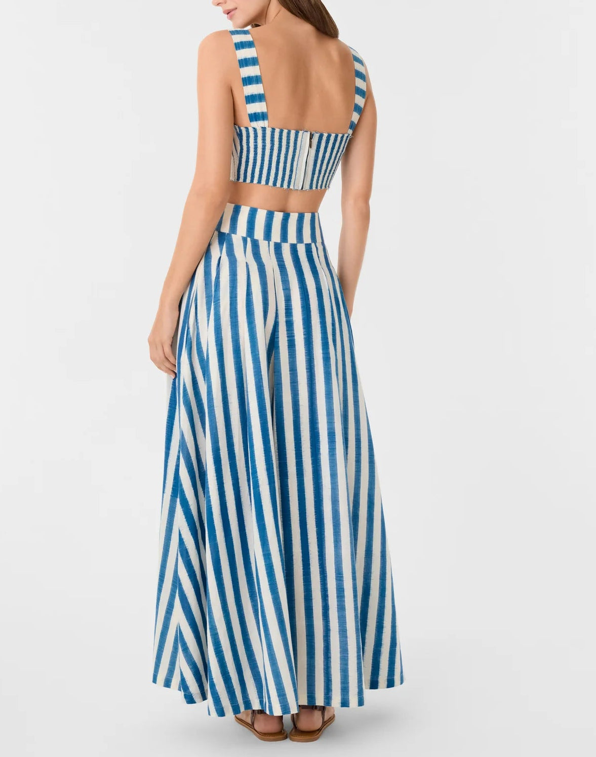 A woman, photographed from the back, wears the Emporio Sirenuse Flaminia Ikat Stripes Skirt—high-waisted, floor-length, in handwoven blue and white stripes—with a matching sleeveless crop top.