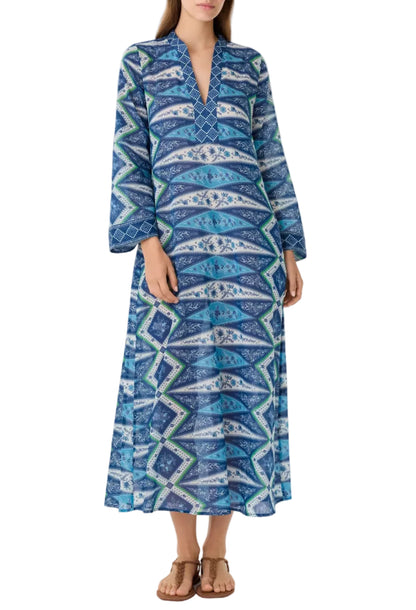 A woman wears the long, loose-fitting blue Jenna Kites Caftan Dress by Emporio Sirenuse, adorned with batik-inspired florals, featuring long sleeves, a V-neckline, and paired with sandals.