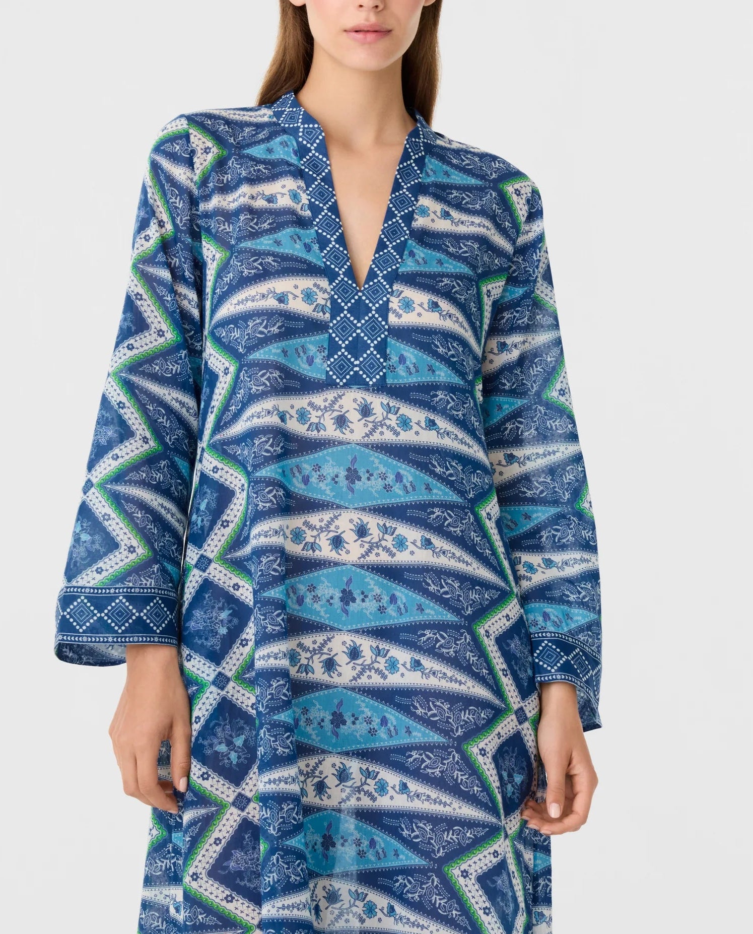 A person wearing the Jenna Kites Caftan Dress by Emporio Sirenuse, a long-sleeved blue dress with a v-neckline, adorned with geometric and batik-inspired floral patterns.