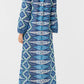 A person stands facing away, wearing the Emporio Sirenuse Jenna Kites Caftan Dress—an ankle-length, long-sleeved dress with batik-inspired blue florals—paired with brown sandals against a plain light background.