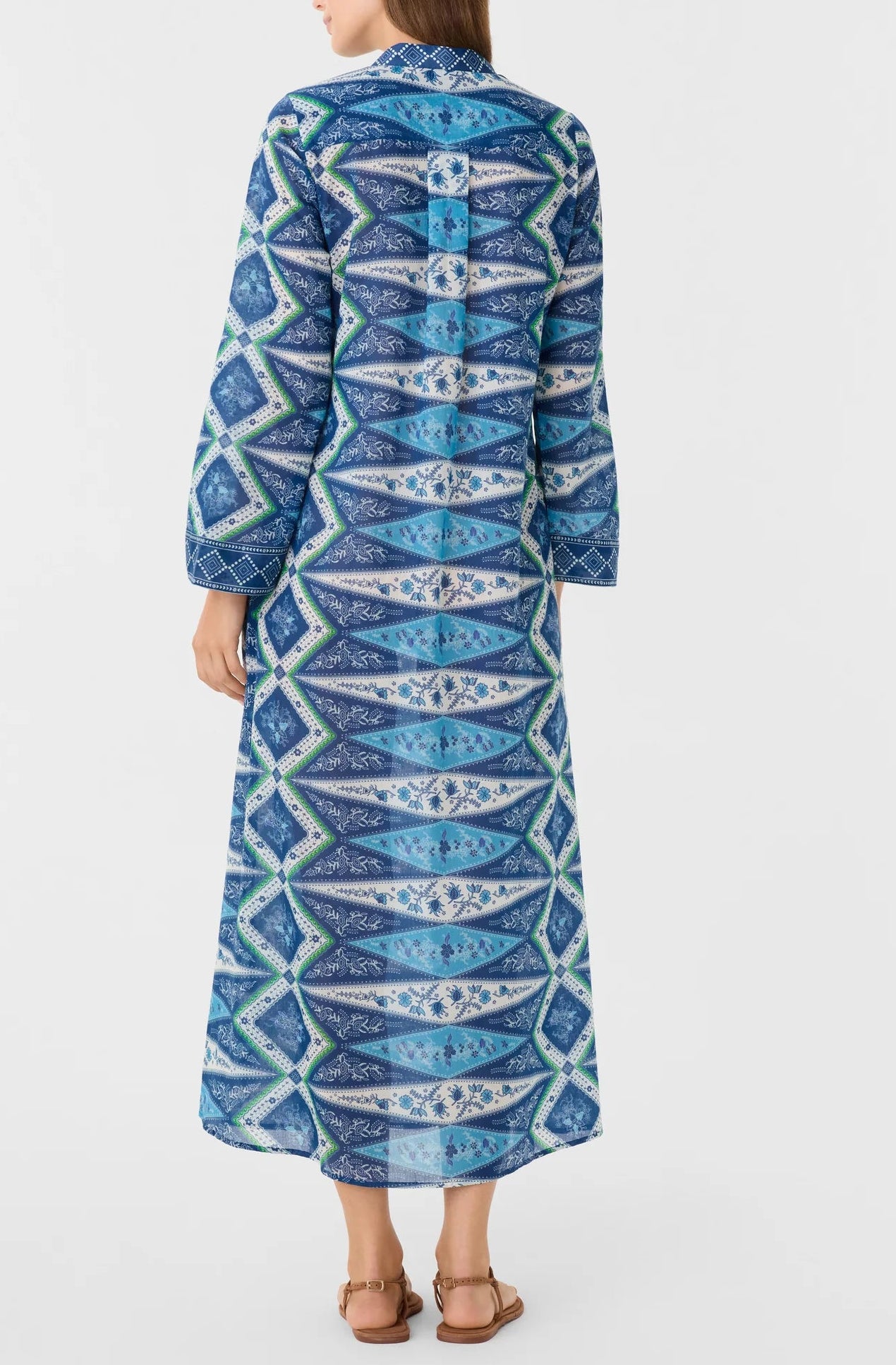 A person stands facing away, wearing the Emporio Sirenuse Jenna Kites Caftan Dress—an ankle-length, long-sleeved dress with batik-inspired blue florals—paired with brown sandals against a plain light background.