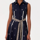 A woman wears the Emporio Sirenuse Miralda Dress with Iris Embroidery, a sleeveless, collared navy cotton poplin dress featuring white geometric patterns and a patterned beige belt at the waist.