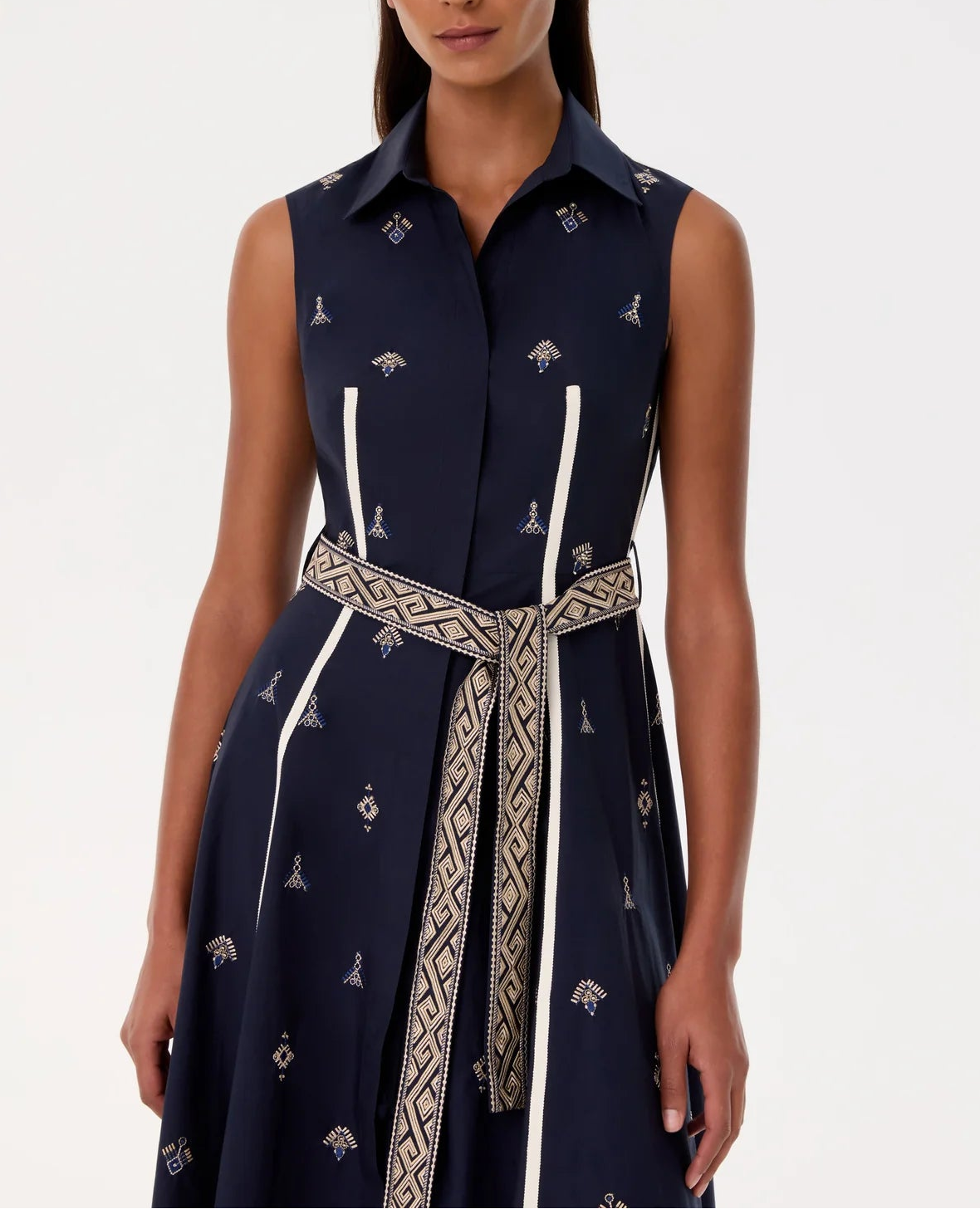 A woman wears the Emporio Sirenuse Miralda Dress with Iris Embroidery, a sleeveless, collared navy cotton poplin dress featuring white geometric patterns and a patterned beige belt at the waist.