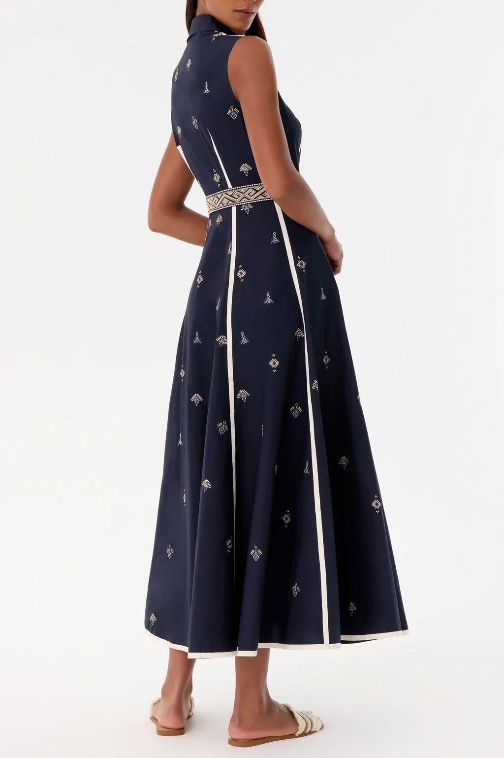 A woman stands in sandals against a plain background, wearing the Emporio Sirenuse Miralda Dress with Iris Embroidery—a sleeveless navy maxi dress featuring white trim and intricate patterned details.