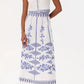 A woman in a sleeveless white dress with blue embroidery and tan sandals exudes effortless chic—her look pairs perfectly with the Emporio Sirenuse Camille Skirt with Iris Embroidery for added style.
