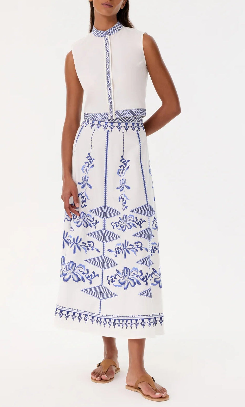 A woman in a sleeveless white dress with blue embroidery and tan sandals exudes effortless chic—her look pairs perfectly with the Emporio Sirenuse Camille Skirt with Iris Embroidery for added style.