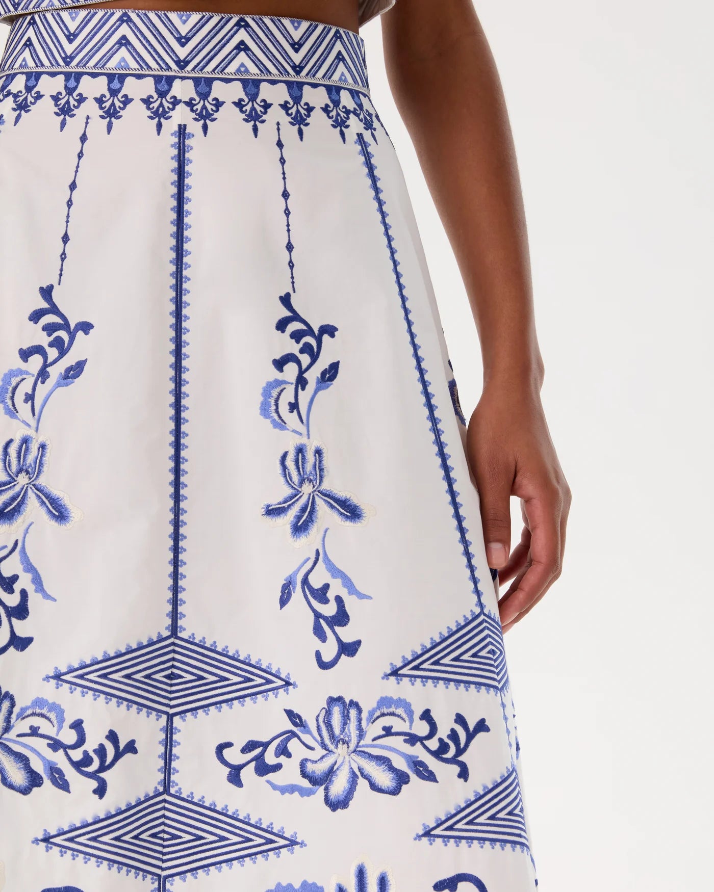 A person models the Emporio Sirenuse Camille Skirt with Iris Embroidery, featuring blue floral and geometric designs on white cotton poplin, shown from waist to mid-thigh against a plain background.