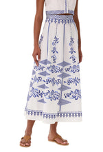 A person wears the Emporio Sirenuse Camille Skirt with Iris Embroidery—a white cotton poplin skirt featuring blue floral and geometric designs—paired with a white top and brown sandals.