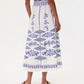 A woman, barefoot and seen from the back against a plain background, wears the Emporio Sirenuse Camille Skirt with Iris Embroidery—a white cotton poplin midi skirt adorned with blue geometric and floral designs.