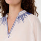 Close-up of a woman wearing the Emporio Sirenuse Ilizia Blouse with Iris Embroidery, featuring a cotton-silk blend, V-shaped neckline, and blue hand-finished embroidery on the collar and shoulders.