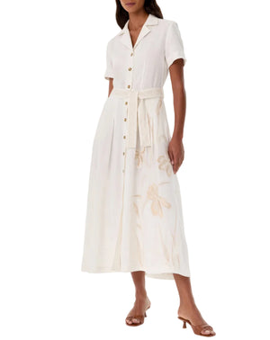 A woman wears the Emporio Sirenuse Daria Sand Flowers Dress, a short-sleeved midi chemisier with a waist tie and beige floral embroidery, paired with brown heeled sandals.