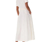 A woman in the Emporio Sirenuse Daria Sand Flowers Dress, a short-sleeve white linen dress with a belted waist, paired with brown heeled sandals, stands with her back to the camera.