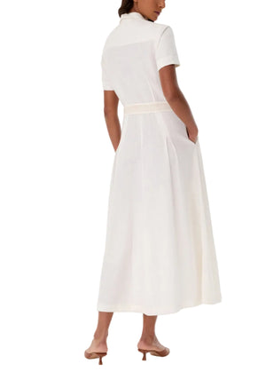 A woman in the Emporio Sirenuse Daria Sand Flowers Dress, a short-sleeve white linen dress with a belted waist, paired with brown heeled sandals, stands with her back to the camera.