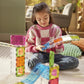 A young girl sits on the floor in the living room, building an imaginative layout with colorful, transparent plastic tiles and ramps from her Magnatiles Rail Racers Max 80-Piece Set.