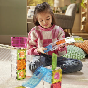 A young girl sits on the floor in the living room, building an imaginative layout with colorful, transparent plastic tiles and ramps from her Magnatiles Rail Racers Max 80-Piece Set.