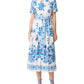 A woman wears the Emporio Sirenuse Claudia Chintz Dress, a white midi with blue botanical print, short sleeves, a tied waist belt, and coordinating blue lace-up sandals.