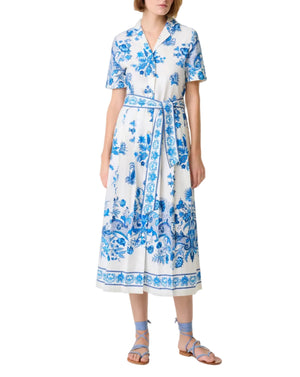 A woman wears the Emporio Sirenuse Claudia Chintz Dress, a white midi with blue botanical print, short sleeves, a tied waist belt, and coordinating blue lace-up sandals.