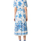 A woman stands facing away, wearing the Emporio Sirenuse Claudia Chintz Dress—a white, short-sleeve midi dress with blue botanical print—paired with matching blue sandals.