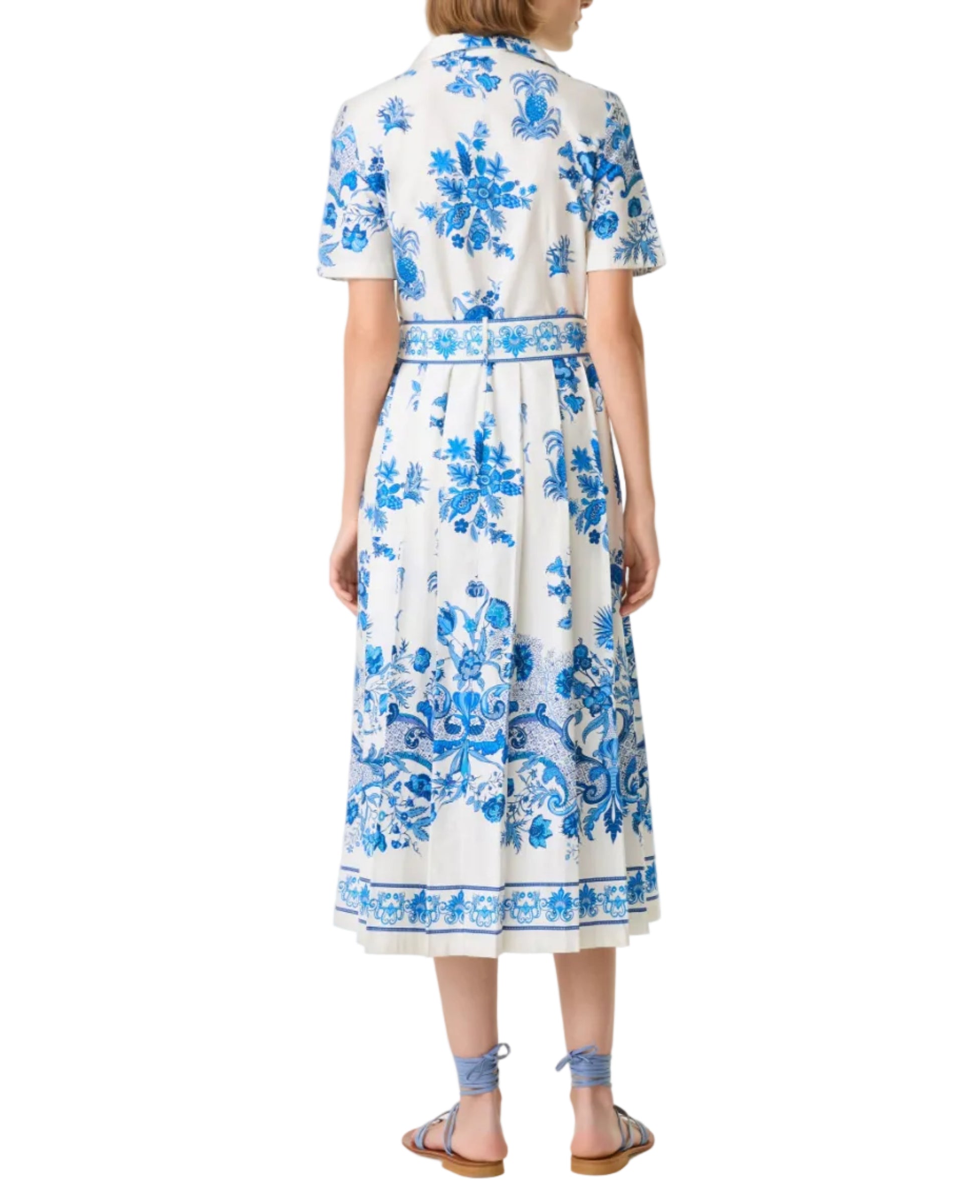 A woman stands facing away, wearing the Emporio Sirenuse Claudia Chintz Dress—a white, short-sleeve midi dress with blue botanical print—paired with matching blue sandals.