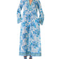 A person wearing the Emporio Sirenuse Jenna Eastern Dream Dress—a long, blue and white cotton voile dress with long sleeves, a patterned neckline, and matching flat sandals.