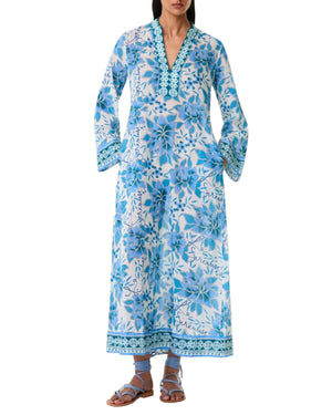 A person wearing the Emporio Sirenuse Jenna Eastern Dream Dress—a long, blue and white cotton voile dress with long sleeves, a patterned neckline, and matching flat sandals.