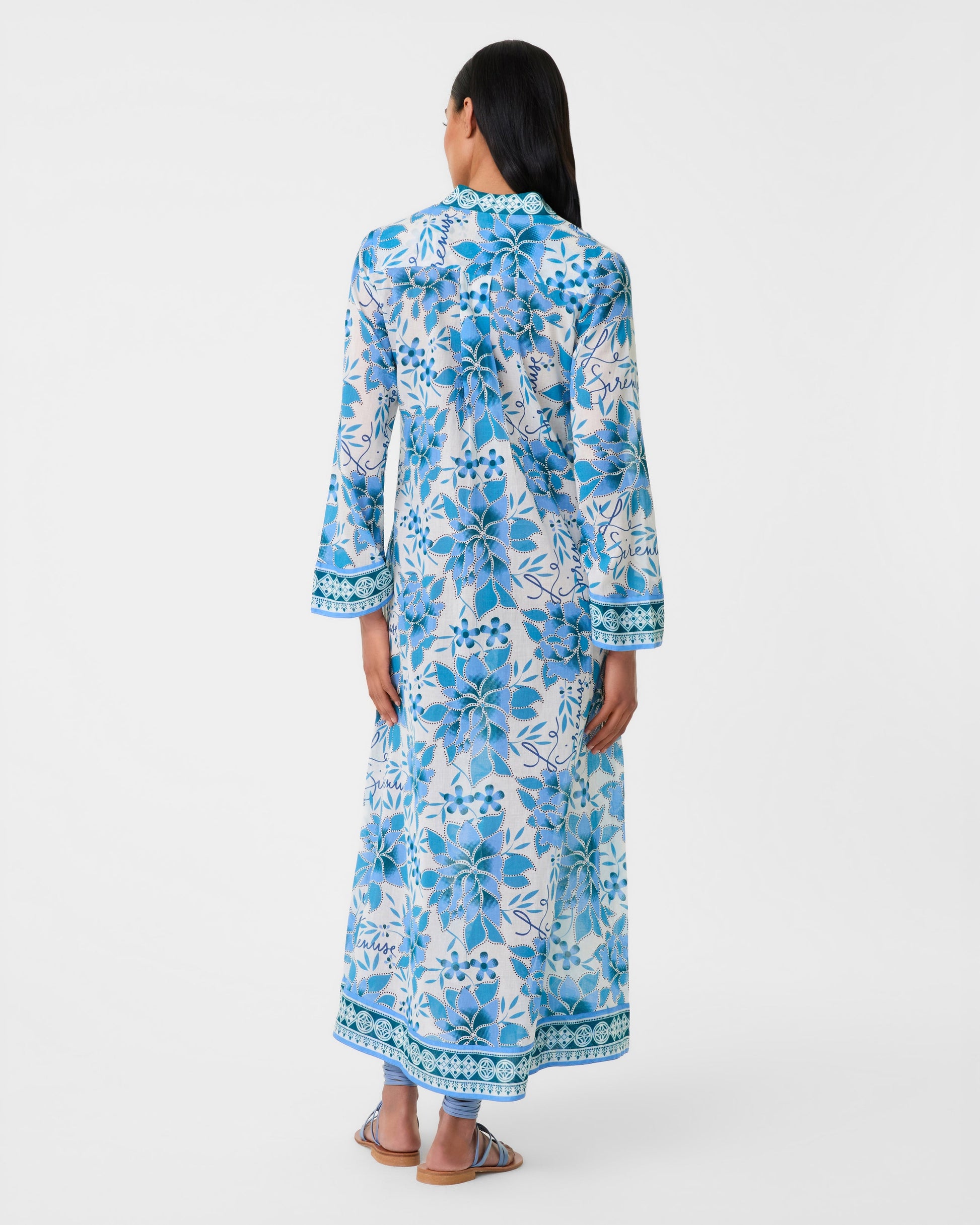 A person stands facing away, wearing the ankle-length Jenna Eastern Dream Dress by Emporio Sirenuse in airy cotton voile, paired with blue sandals.