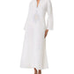 A woman wears the Emporio Sirenuse Jenna Meridiana Dress, a long white linen-cotton blend with long sleeves, V-neck, and Meridiana Embroidery on the chest, paired with brown flat sandals.