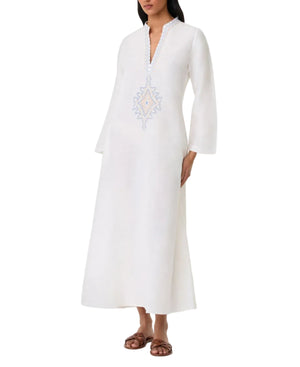 A woman wears the Emporio Sirenuse Jenna Meridiana Dress, a long white linen-cotton blend with long sleeves, V-neck, and Meridiana Embroidery on the chest, paired with brown flat sandals.