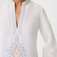 A woman wears the Jenna Meridiana Dress by Emporio Sirenuse—a white long-sleeve linen-cotton dress with a zigzag embroidered V-neck and blue and beige Meridiana Embroidery on the chest.