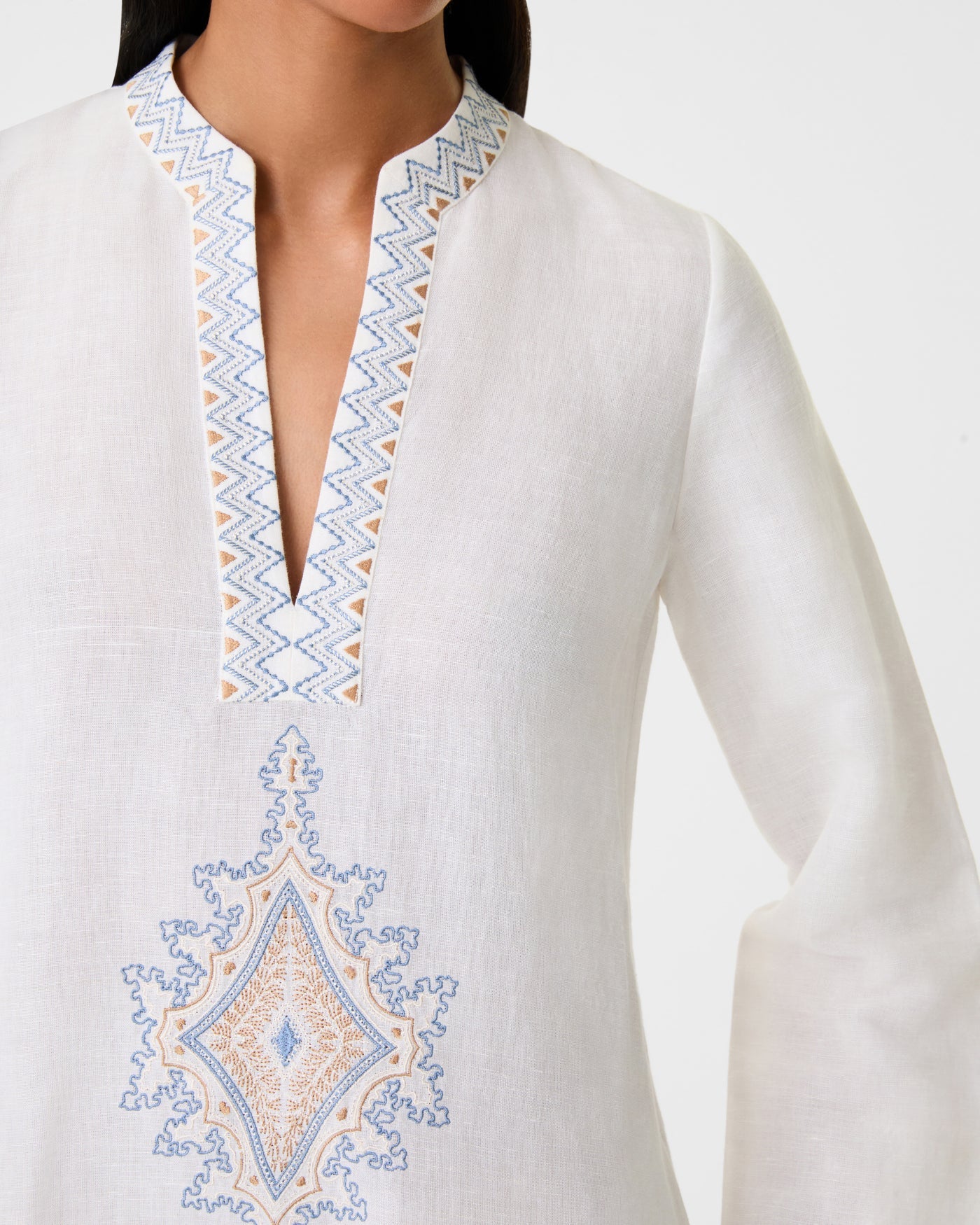 A woman wears the Jenna Meridiana Dress by Emporio Sirenuse—a white long-sleeve linen-cotton dress with a zigzag embroidered V-neck and blue and beige Meridiana Embroidery on the chest.