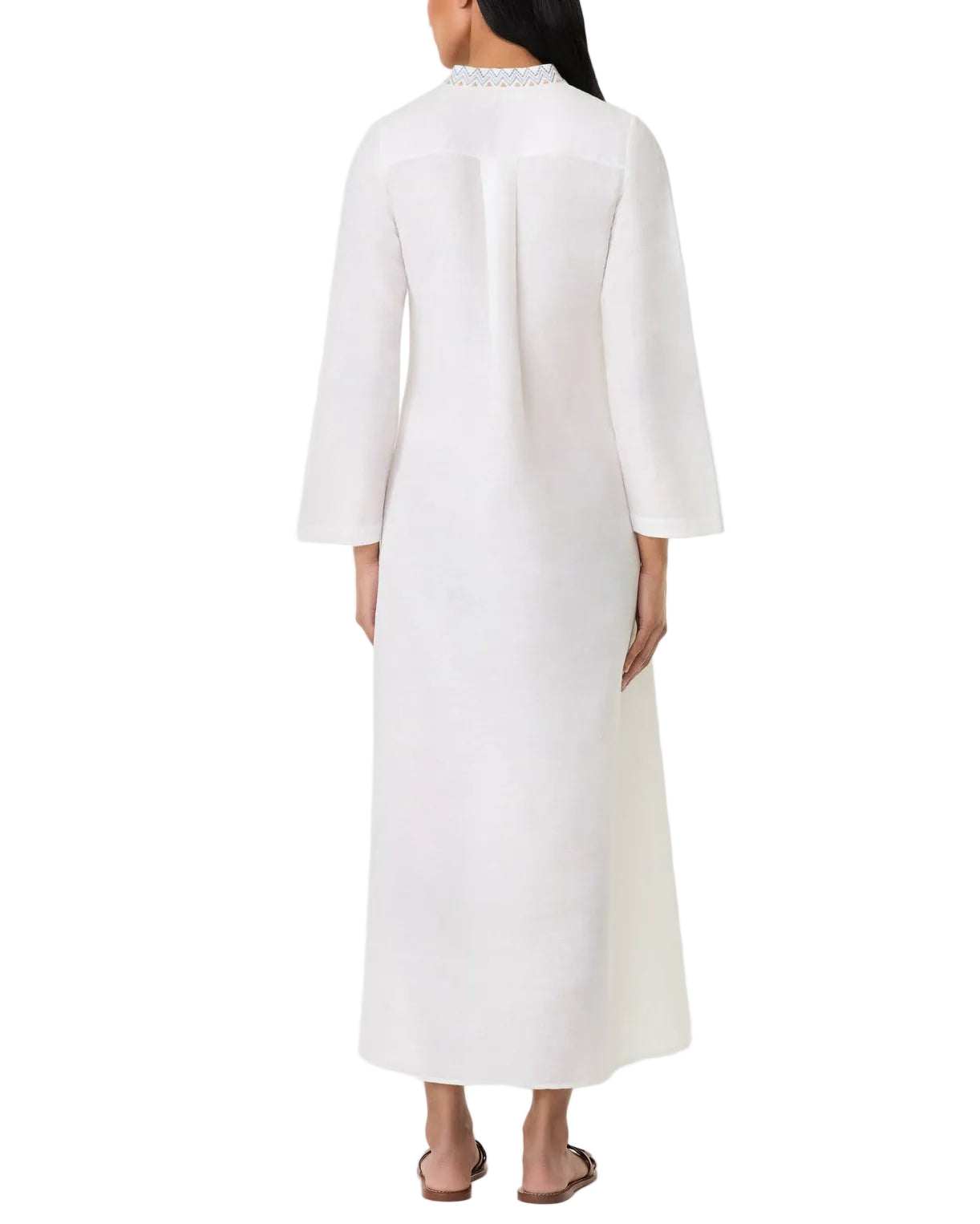 A woman stands facing away, wearing the Jenna Meridiana Dress by Emporio Sirenuse—an ankle-length, long-sleeved white dress with Meridiana embroidery and a loose, graceful fit.