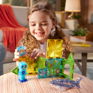A young child plays with the MAGNATILES Shark Dive 13-Piece Set, which features colorful magnetic tiles, a blue figurine, and a toy shark on a table in the living room, creating an imaginative underwater play scene.