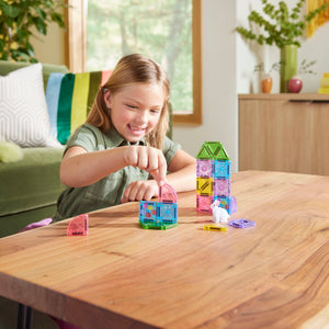 A young girl builds colorful geometric structures with the translucent MicroMags Spring Garden 24-Piece Travel Set by Magnatiles on a wooden table in a bright living room.
