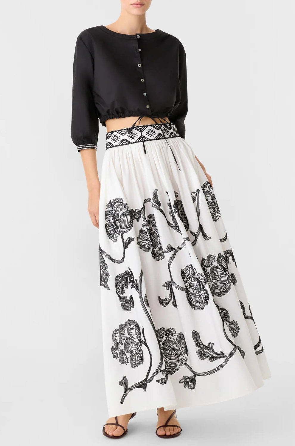 A person wears a black cropped button-up shirt and an Emporio Sirenuse Ombretta Peony Embroidery Skirt in white cotton poplin with black peony embroidery and a patterned waistband.