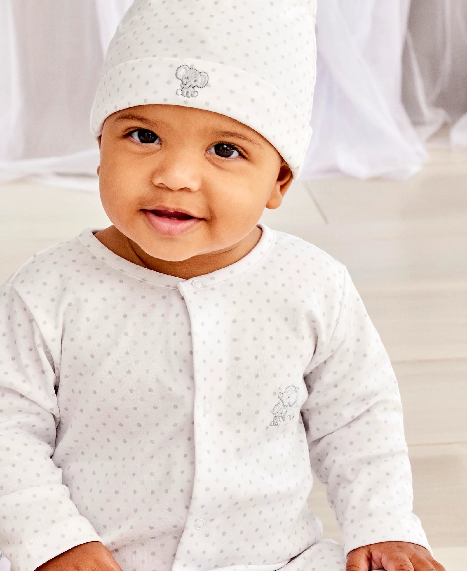 A baby in the Kissy Kissy Baby Jungle Playdate Dot Footie—a white Pima cotton outfit with grey dots and an elephant motif—sits indoors, gazing at the camera and evoking the charm of jungle animals.