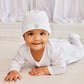 A baby wearing the Kissy Kissy Baby Jungle Playdate Dot Hat smiles at the camera while lying on their stomach on a light-colored floor, with white drapes in the background.