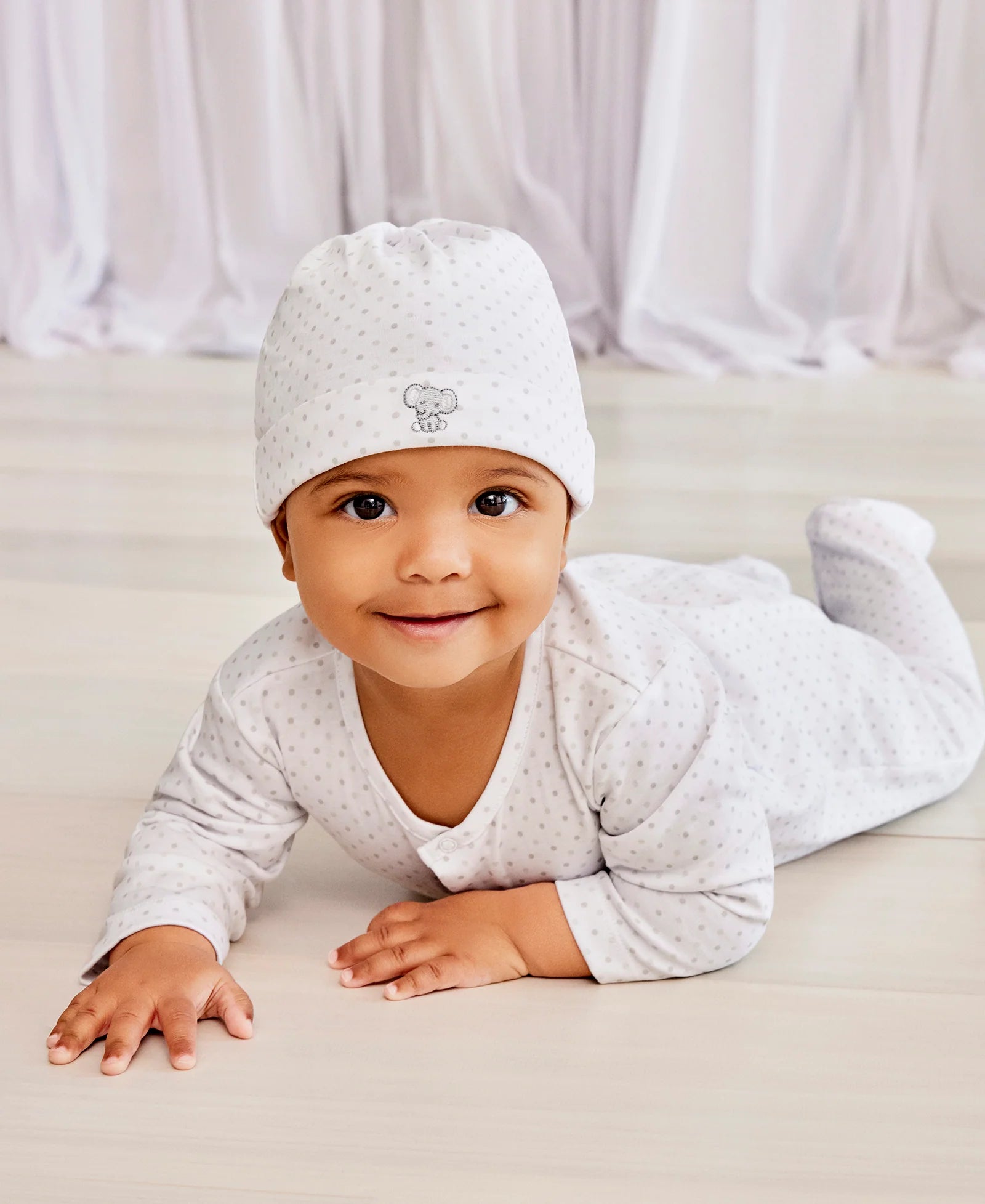A baby wearing the Kissy Kissy Baby Jungle Playdate Dot Hat smiles at the camera while lying on their stomach on a light-colored floor, with white drapes in the background.