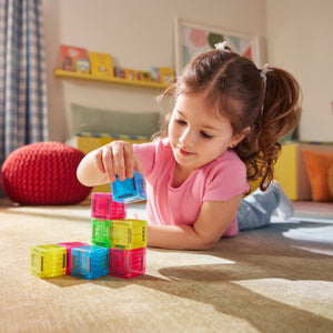 A young child in a pink shirt lies on the floor, stacking colorful MicroMags Cubes 9-Piece Set by Magnatiles in a bright, cozy room.