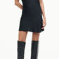 A person models the Staud Iris Silk Dress—a black mini dress with a cowl neck and short sleeves—paired with knee-high black heeled boots against a plain background.