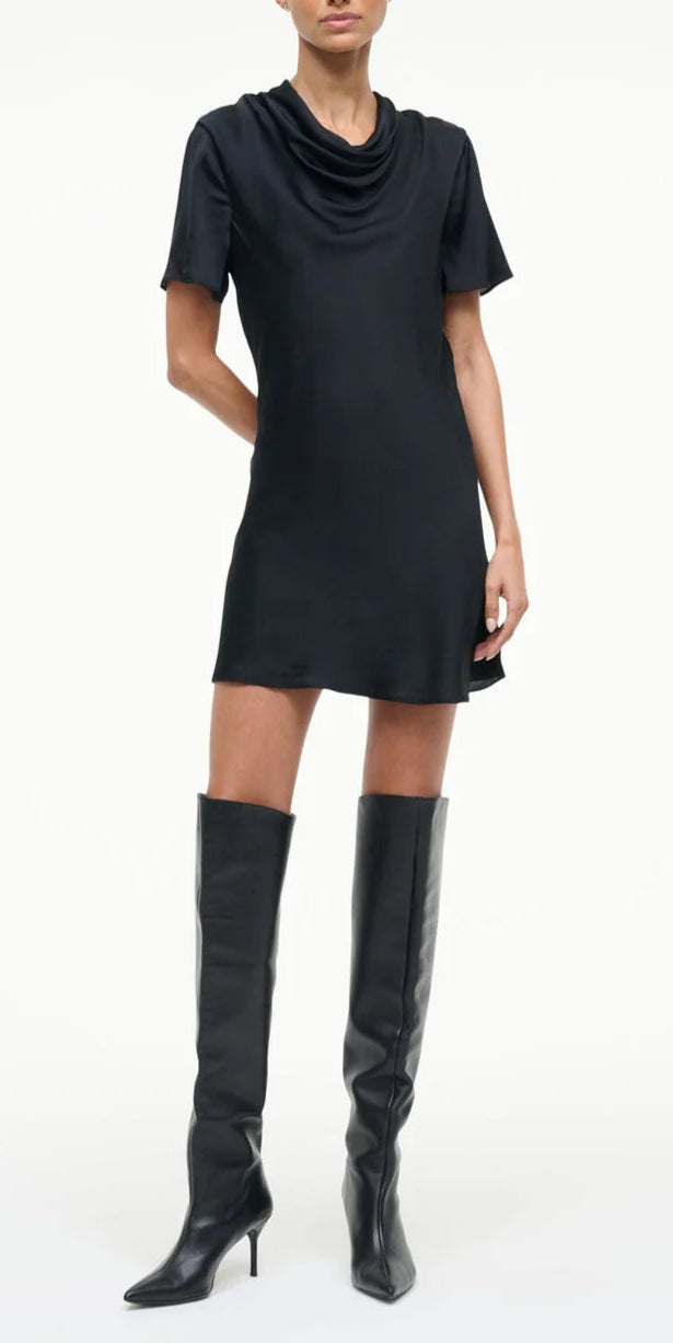 A person models the Staud Iris Silk Dress—a black mini dress with a cowl neck and short sleeves—paired with knee-high black heeled boots against a plain background.
