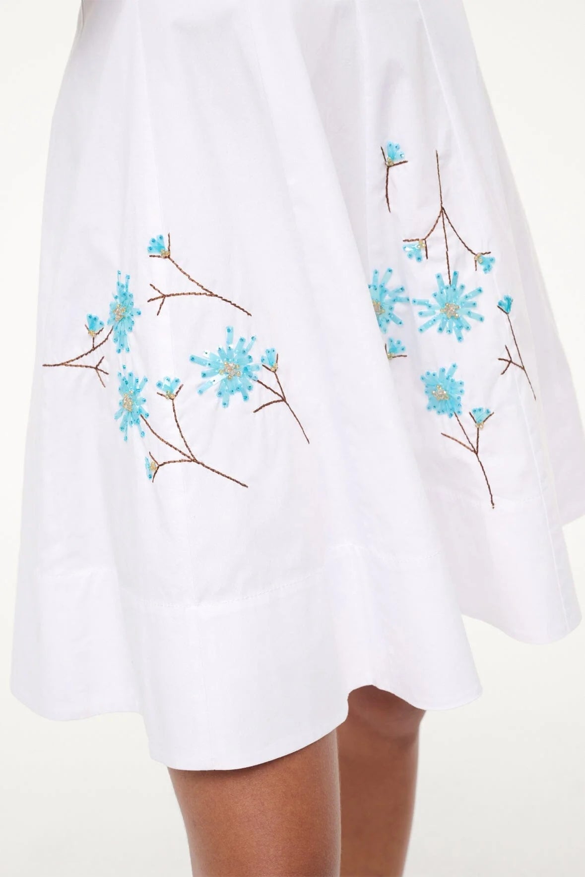 The Staud Mini Wells Dandelion Dress is shown on a model with medium brown skin. This white cotton poplin dress features blue and brown floral embroidery near the hem.