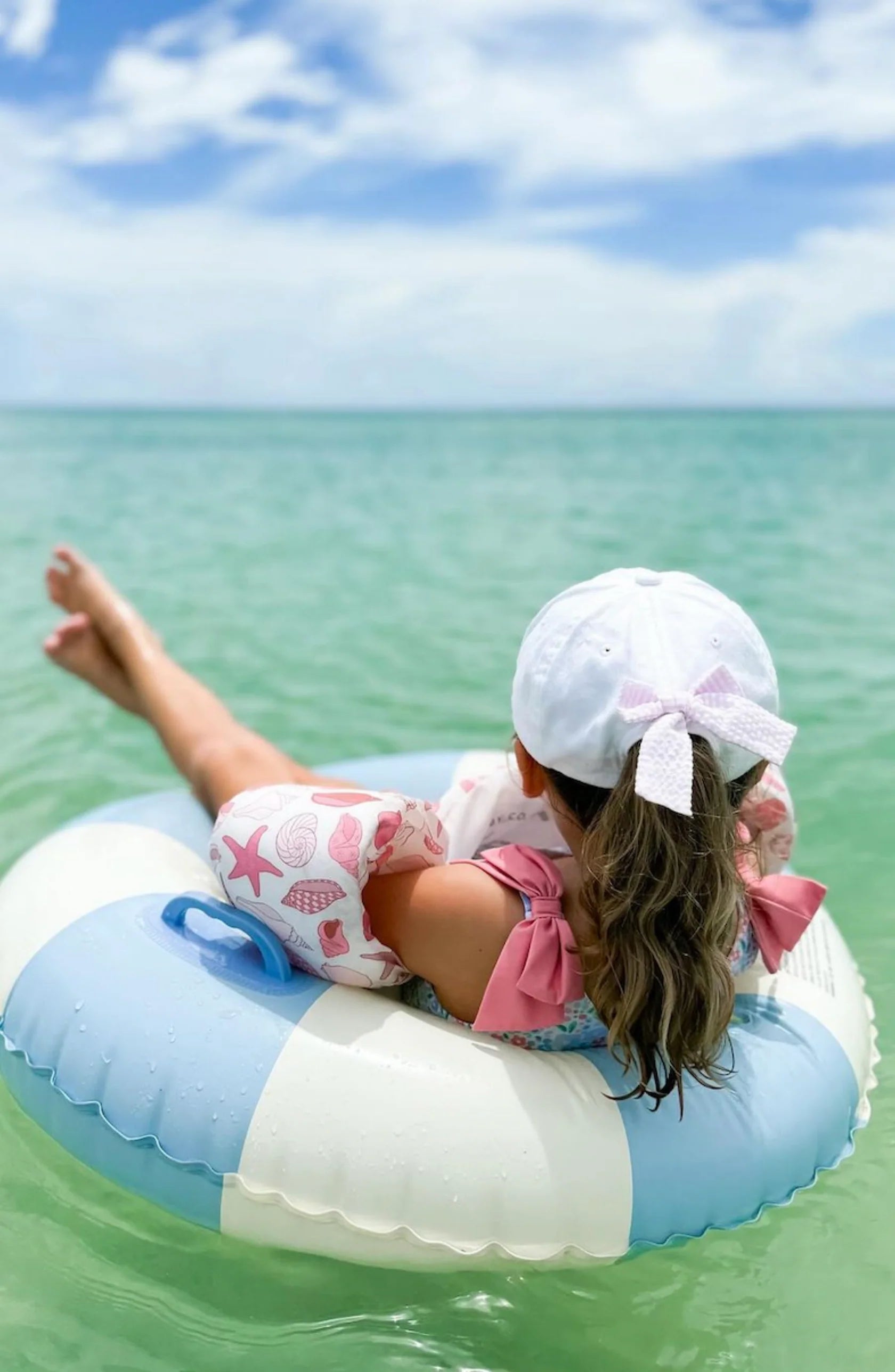 A child wearing the Bits & Bows Birthday Girl Bow Baseball Hat and pink floaties relaxes on a blue and white inflatable ring in calm, clear green ocean water under a partly cloudy sky.